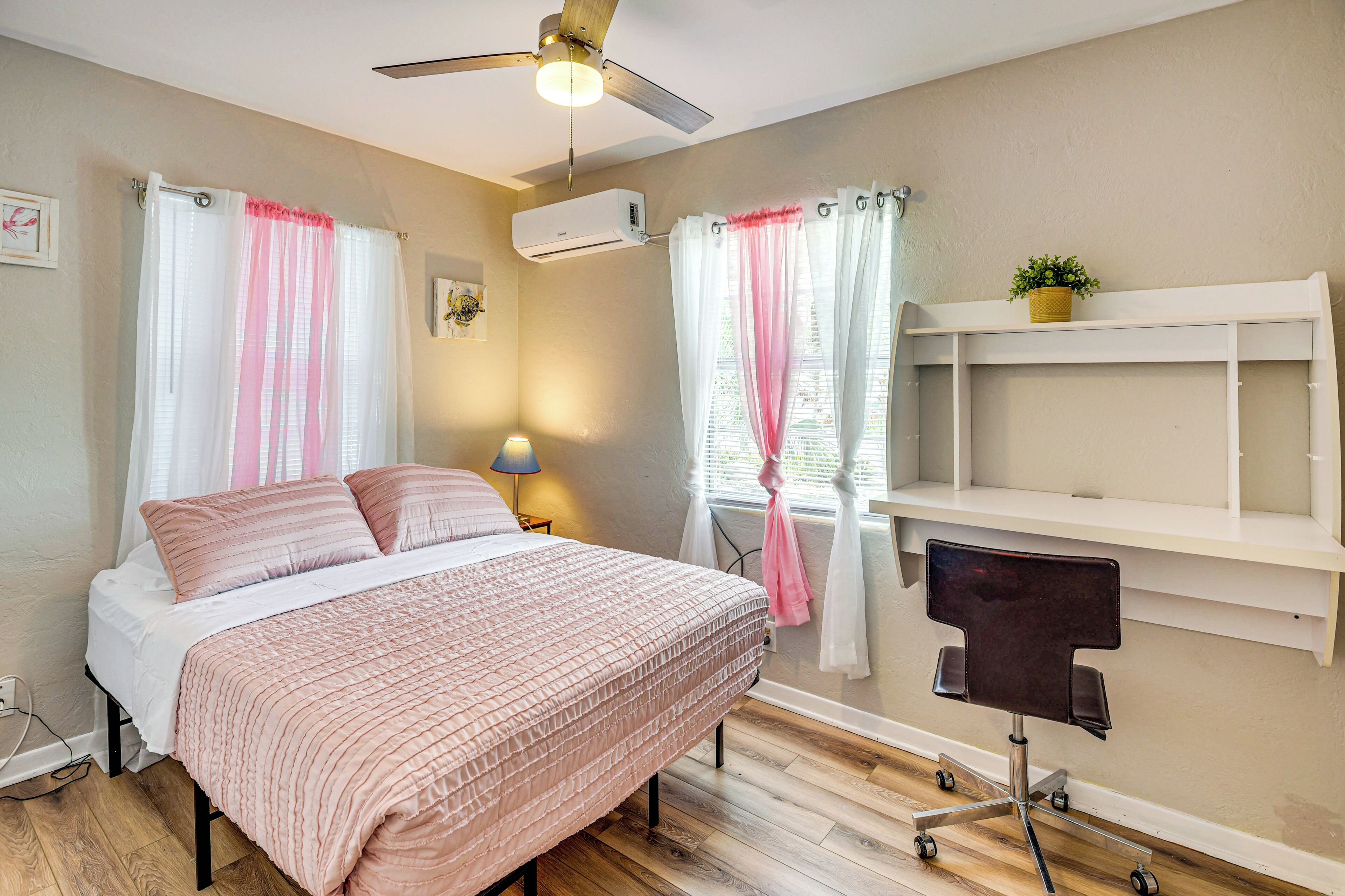 A bedroom with a bed, a ceiling fan, an air conditioner, a desk with a chair, and a window with curtains.