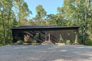A modern cabin with a dark exterior, a front door, and a gravel driveway.
