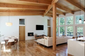 Ein großzügiger Wohnbereich mit Holzbalkendecke, Kamin, Fernseher und großen Fenstern mit Blick ins Freie.