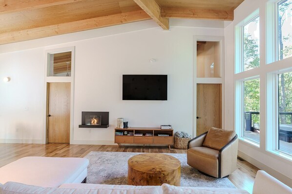 A spacious living room with a fireplace, a large TV, and a view of the outdoors through large windows.