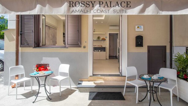 Exterior del Palacio Rossy, que presenta un área de estar al aire libre techada con dos mesas y sillas, una vista hacia el interior y un letrero que indica la ubicación como Suite Costa de Amalfi.