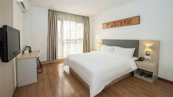 A hotel room with a bed, bedside table, lamp, TV, desk, chair, and curtains.