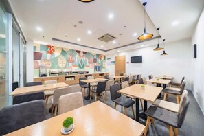 Moderni ruokailutila, jossa on useita pöytiä ja tuoleja, taustalla keittiö ja seinällä värikäs seinämaalaus.