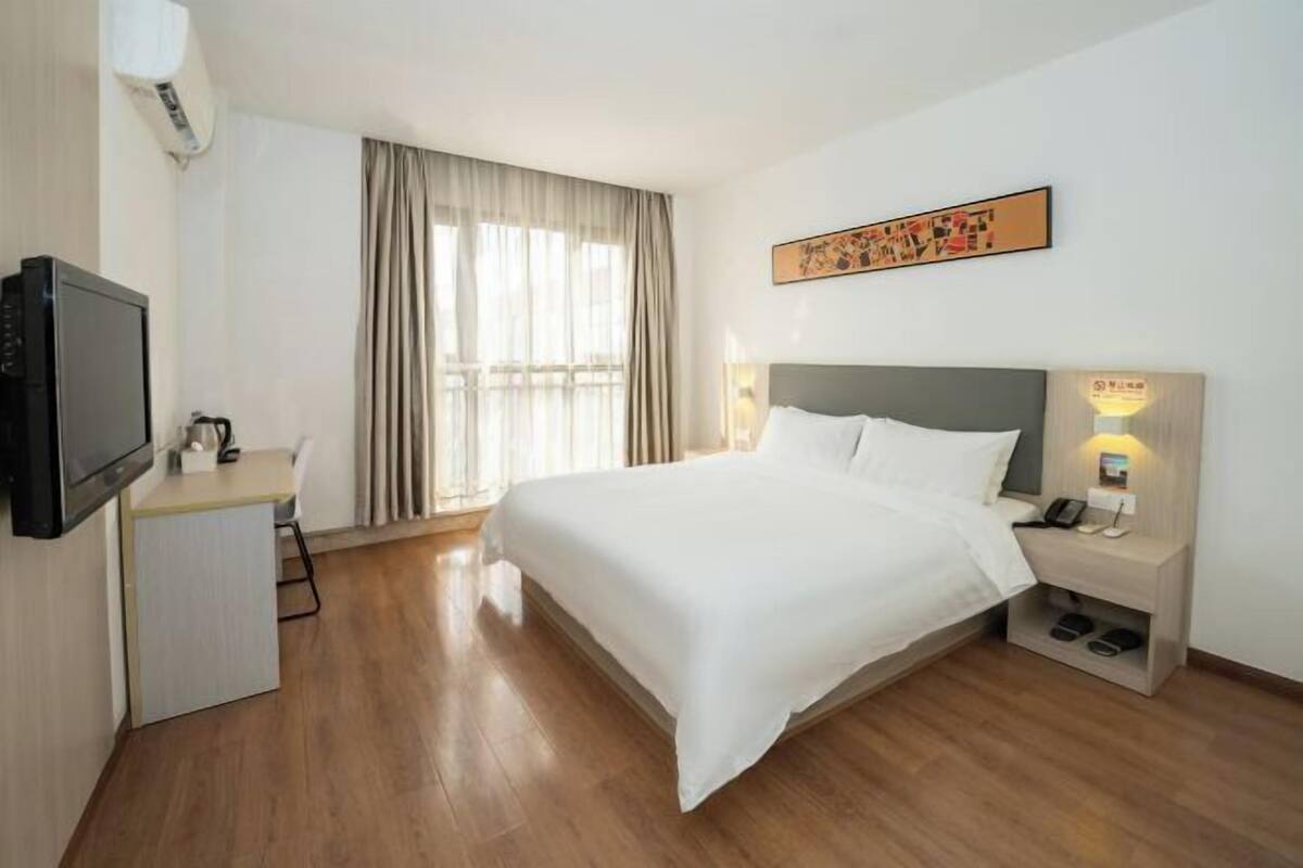 A hotel room with a bed, bedside table, lamp, TV, desk, and curtains.