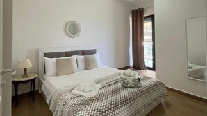A neatly made bed with a patterned blanket, a bedside table with a lamp, and a window with curtains. - FAMILY INN Apartments (Sarande)