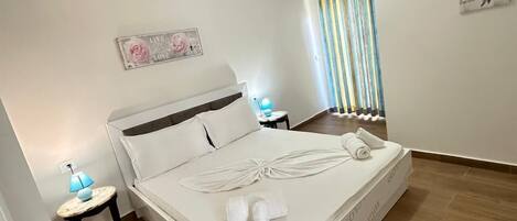 A neatly made bed with a white mattress and pillows, a bedside table with a lamp, and a window with striped curtains.