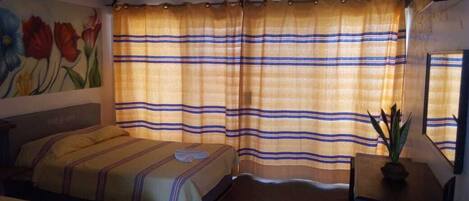 A hotel room with a bed, a small table with a plant, and a window with striped curtains.