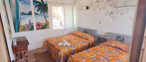 A room with two beds, a ceiling fan, a painting of a beach scene, and a window with curtains.