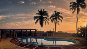 Une piscine entourée d’une terrasse en briques, de palmiers et offrant une vue sur l’océan au coucher du soleil.