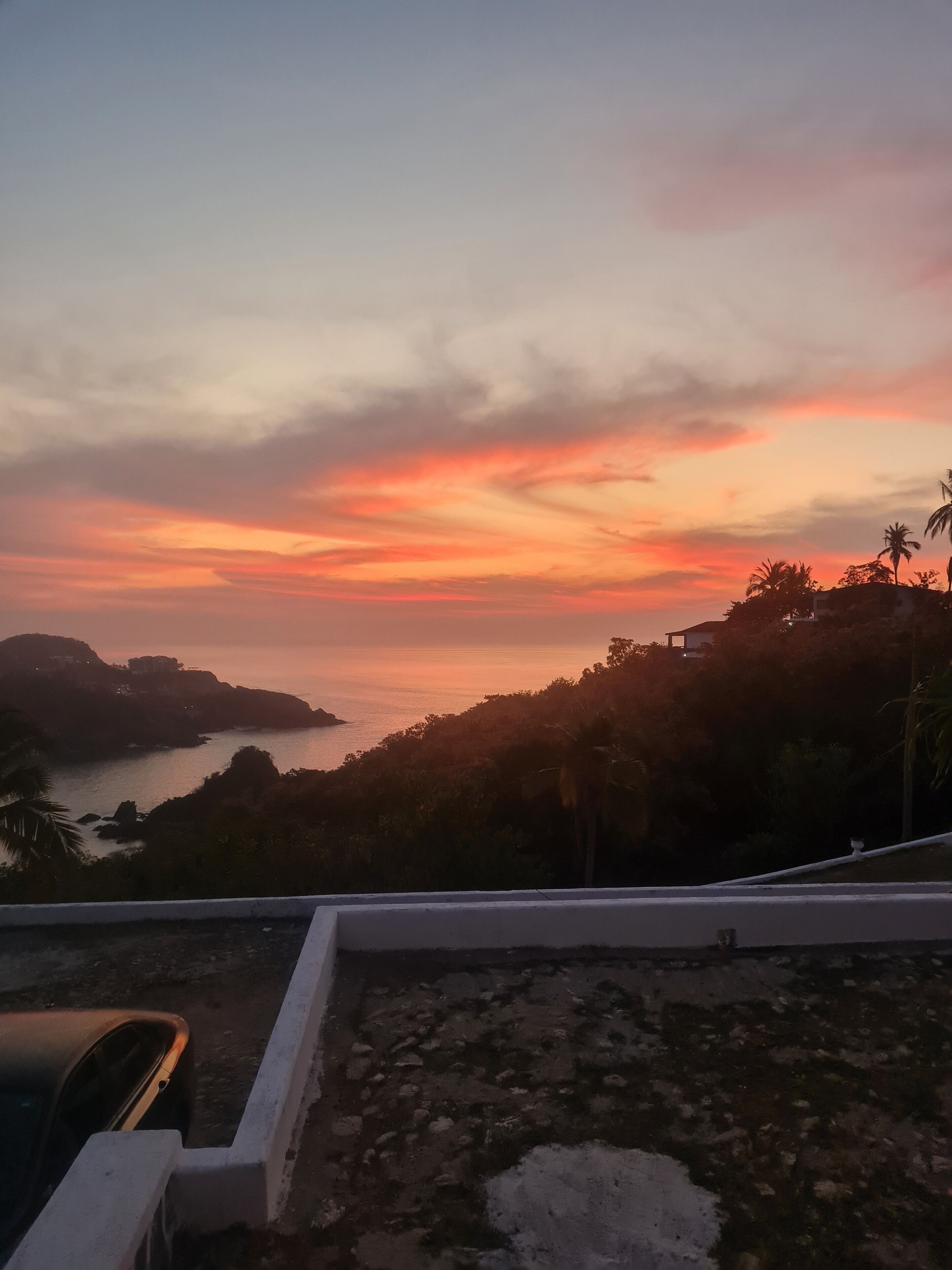 Une vue sur le coucher de soleil depuis un balcon donnant sur une zone côtière, avec des palmiers et une île au loin.