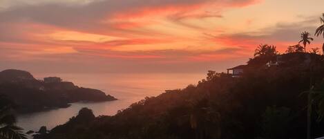 Vista al atardecer desde un balcón con vistas a la costa, palmeras y una isla distante.
