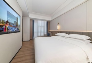 A hotel room with a large bed, a TV screen, a desk, and a window with curtains.
