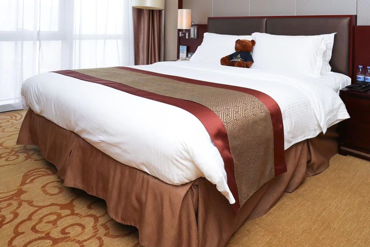 A hotel room with a large bed, a brown bed skirt, a white comforter, and a brown decorative runner. There is a teddy bear on the bed and a nightstand with a lamp and water bottles.