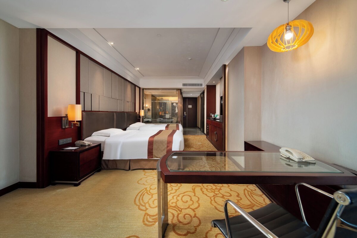 A hotel room with a bed, bedside table, lamp, and a glass partition separating the room from the hallway.