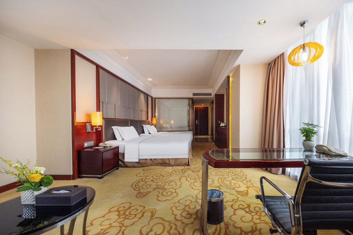 A hotel room with a bed, bedside tables, a desk, a chair, a glass table, a lamp, a potted plant, and a trash can.
