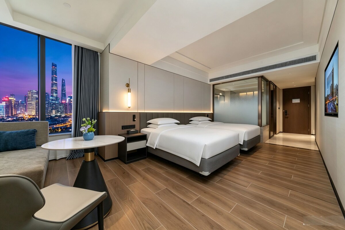 A modern hotel room with a city view, a sofa, a desk, and two beds.