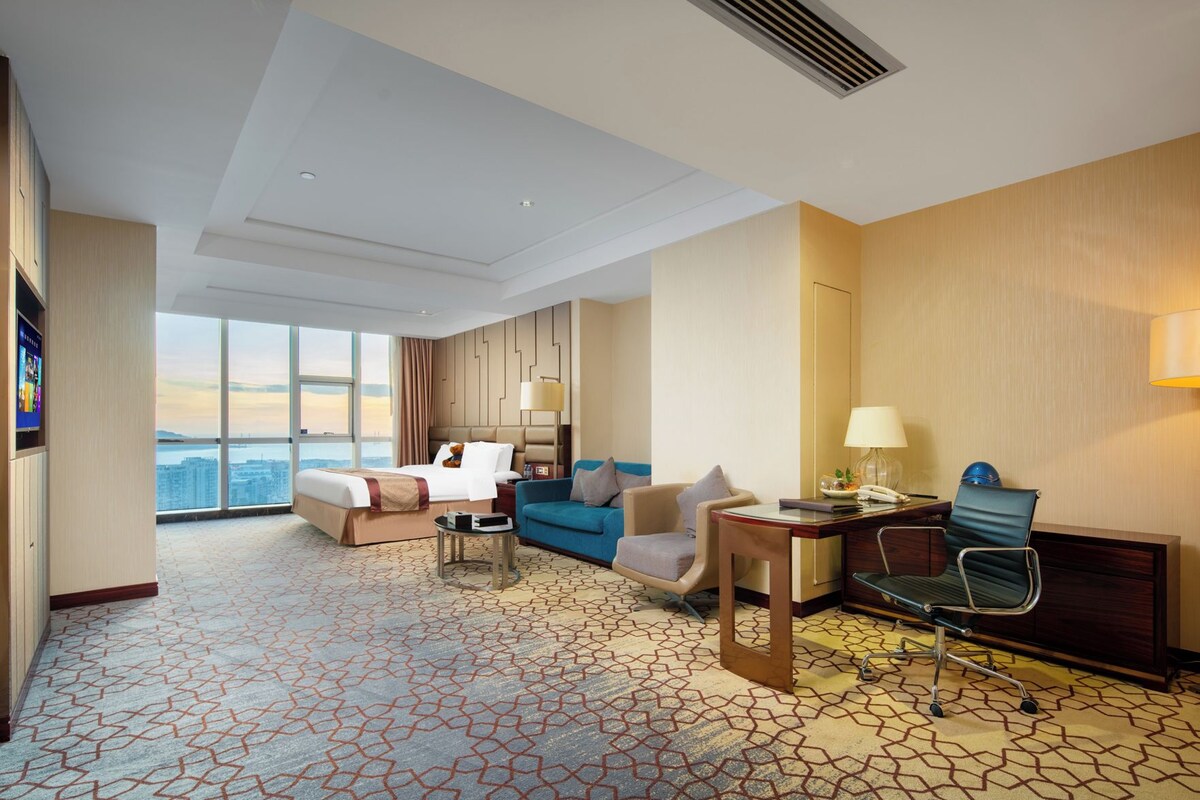 A hotel room with a large bed, a sofa, a desk, and a chair. There is a view of the city through large windows.