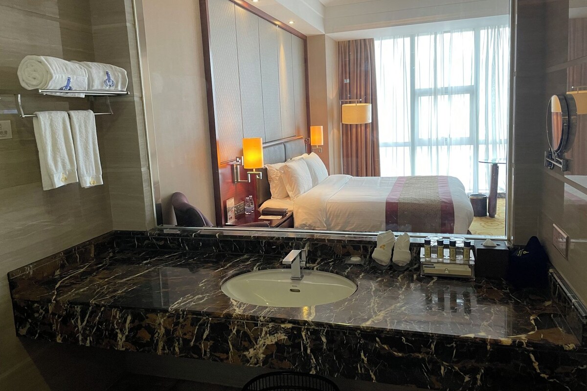 A hotel bathroom with a marble sink, a large mirror, and a bed visible through the glass partition.