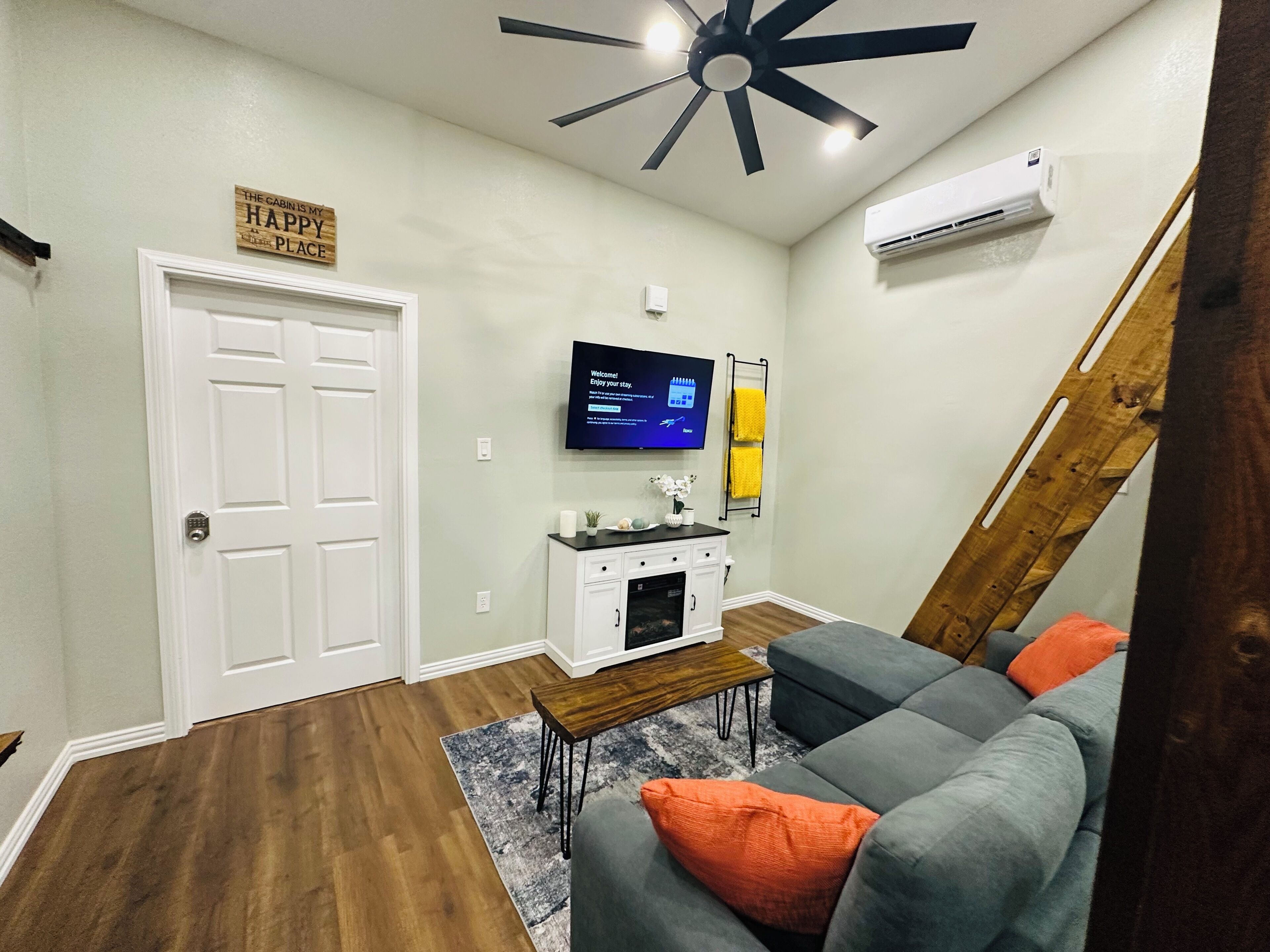 A modern living room with a grey couch, wooden flooring, a ceiling fan, a flat-screen TV, and a sign that says "The Cabin is My Happy Place."