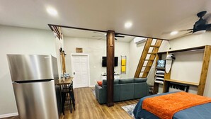 Ein kompakter, moderner Raum mit einer Küchenzeile, einem Bett mit orangefarbener Decke, einem grauen Sofa und einer Holzleiter.