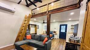 Ein modernes Loft-Zimmer mit Schlafgalerie, grauem Ecksofa, Holztisch und Treppe.