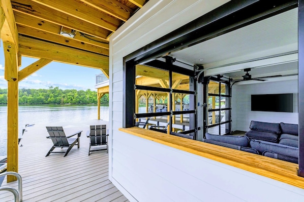 A covered deck with a view of a lake, a seating area with a sofa and chairs, and a large window.