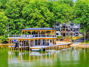 A lakeside wooden structure with a covered deck, a dock extending into the water, and a modern building in the background.