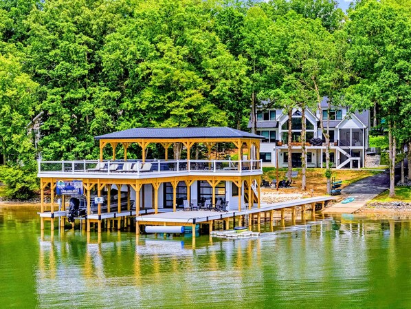 A lakeside wooden structure with a covered deck, a dock extending into the water, and a modern building in the background.