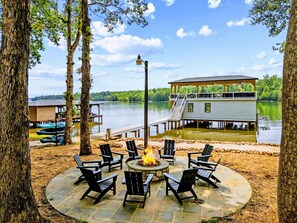 A lakeside deck with outdoor seating and a fire pit, overlooking a dock and a house.