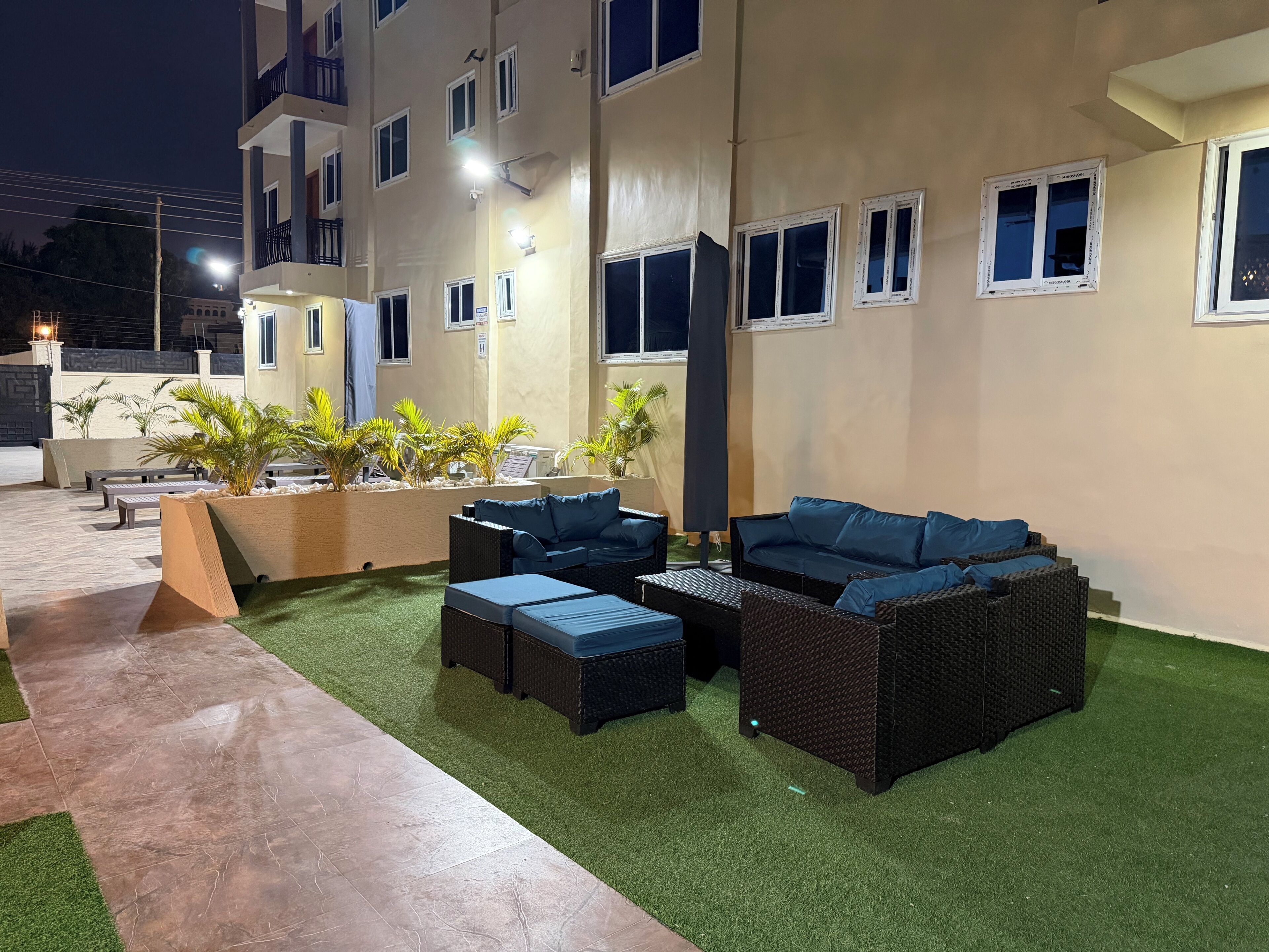A well-lit outdoor seating area with wicker furniture and artificial grass, adjacent to a building with multiple windows and balconies.