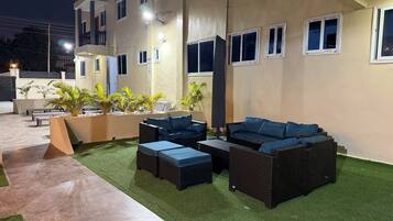 A well-lit outdoor seating area with wicker furniture and artificial grass, adjacent to a building with multiple windows and balconies.