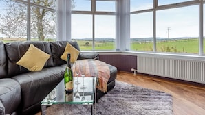 A cozy living room with a leather sofa, a glass coffee table, and a view of a green field through large windows. - 5 bedroom accommodation in Inverurie (Inverurie)