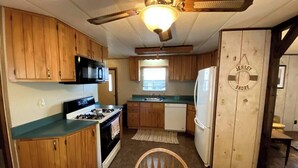 A compact kitchen with wooden cabinets, a stove, a refrigerator, and a ceiling fan. - Escape to Comfort Near Higgins Lake! (Roscommon)