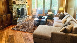 A cozy living room with a stone fireplace, wooden flooring, and a large window offering a view of greenery. - Luxury Lodge Retreat in the Heart of Suncadia – Walk to Golf, Winery & Trails (Cle Elum)