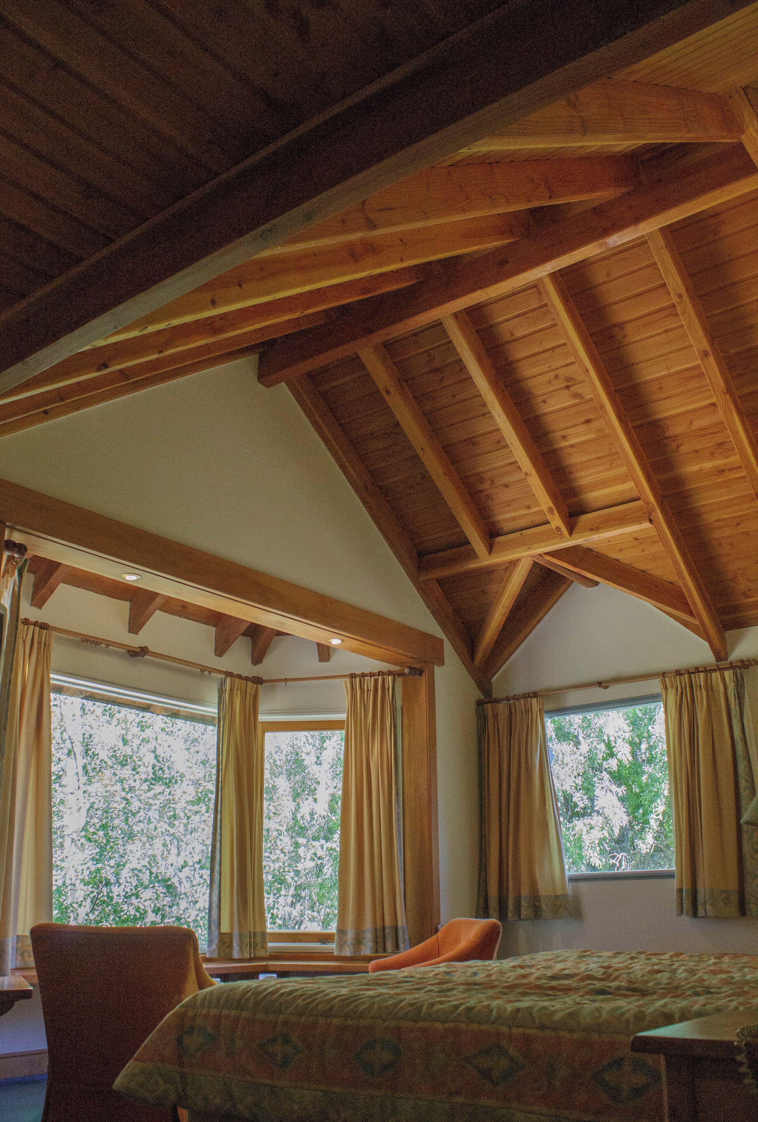 A room with a sloped ceiling, wooden beams, a bed, a chair, and windows with curtains.