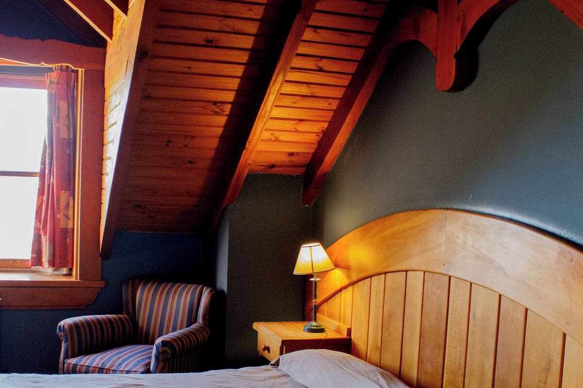 A bedroom with a wooden headboard, a striped armchair, a bedside table with a lamp, and a window with curtains.