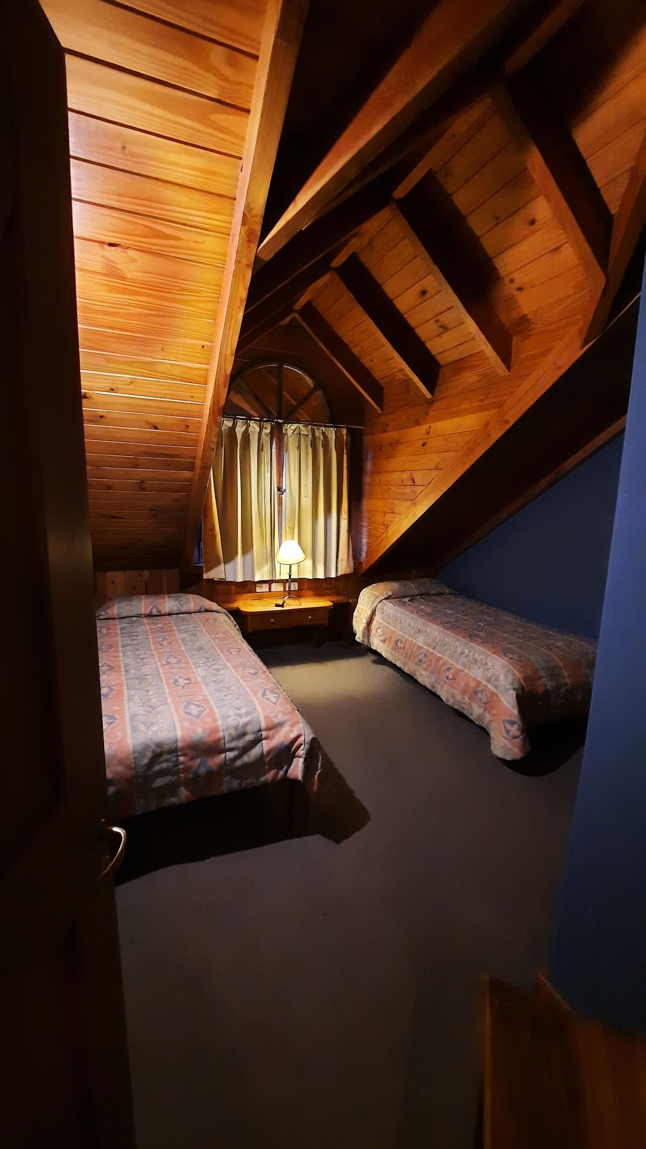 A small attic bedroom with two beds, a window with curtains, and a lamp.