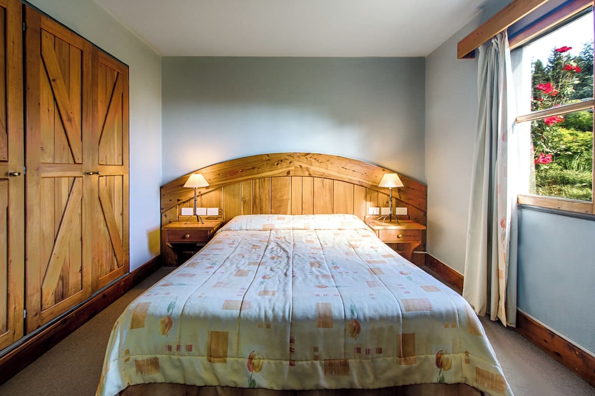 A bedroom with a wooden headboard, two bedside tables with lamps, a bed with a floral-patterned bedspread, and a view of greenery through a window.