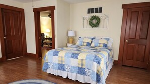 A bedroom with a large bed, wooden flooring, a nightstand, a lamp, and a wall-mounted wreath.