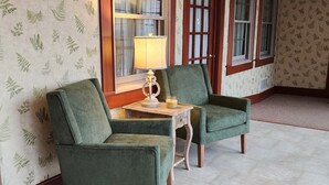 A room with two green armchairs, a small table with a lamp, and large windows with wooden frames.