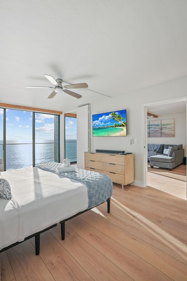 A modern bedroom with a large bed, a ceiling fan, and a view of the ocean through large windows.