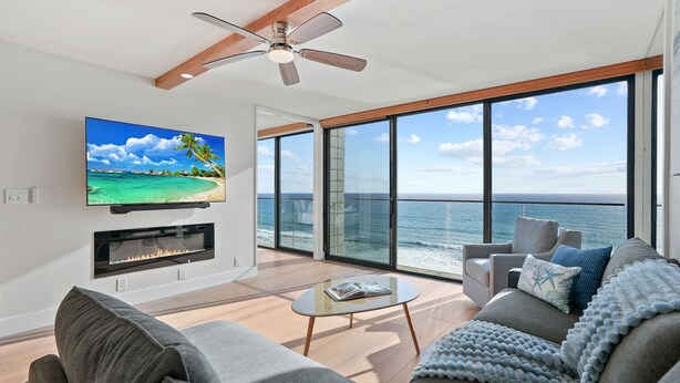A modern living room with a large TV, fireplace, and a balcony with a view of the ocean. - Oceanfront Paradise with Sunset Balcony, Pool, Spa, Beach Access & Parking (San Diego)