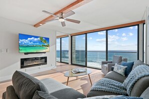 A modern living room with a large TV, fireplace, and a balcony with a view of the ocean.