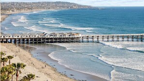 A beach with a pier extending into the ocean, a sandy shore, and a distant cityscape. - Oceanfront Paradise with Sunset Balcony, Pool, Spa, Beach Access & Parking (San Diego)