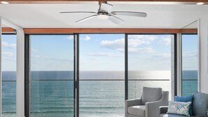 A modern room with a large sliding glass door leading to a balcony with a view of the ocean. There is a ceiling fan, a round table with a magazine, and two grey armchairs. - Oceanfront Paradise with Sunset Balcony, Pool, Spa, Beach Access & Parking (San Diego)