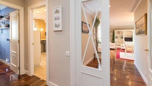 Un pasillo con suelo de madera, puertas blancas y vista a un comedor con alfombra roja y mobiliario blanco.
