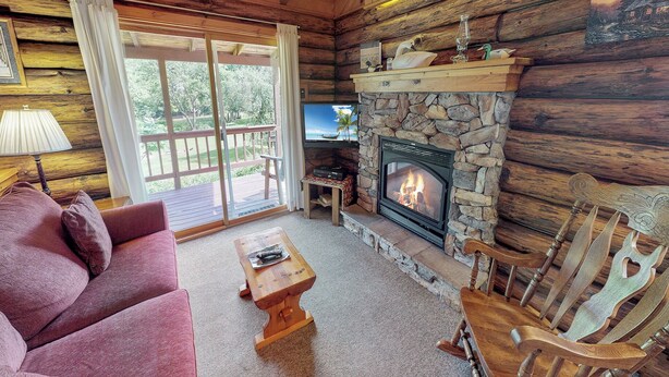 Ein gemütliches Blockhaus-Interieur mit Kamin aus Stein, einem darüber montierten Fernseher und einem Holzschaukelstuhl.