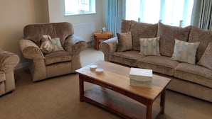 A living room with a brown sofa, a brown armchair, and a wooden coffee table. There is a window with curtains and a radiator. - Pen Y Frith Bungalow (Prestatyn)