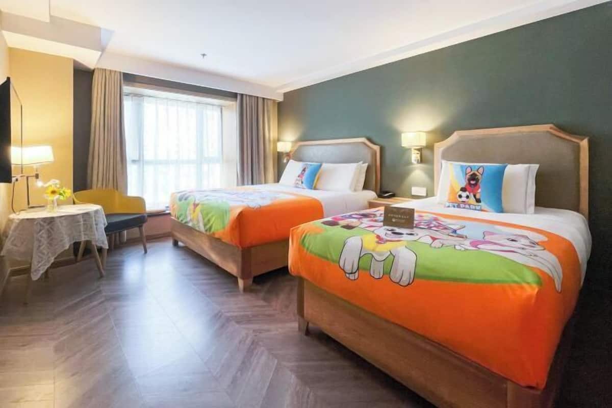 A hotel room with two beds, a small table with a chair, and a window with curtains.