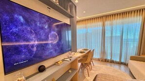 A modern living room with a large flat-screen TV displaying a space scene, a dining area with a table set for two, and a cozy seating area with a bed. - RN Studio Premium 512 - Frente Mar (Maceio)
